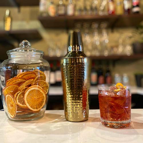 Red Alcoholic Drink on Bar Counter beside Dried Orange Slices in Glass Jar and Silver Shaker